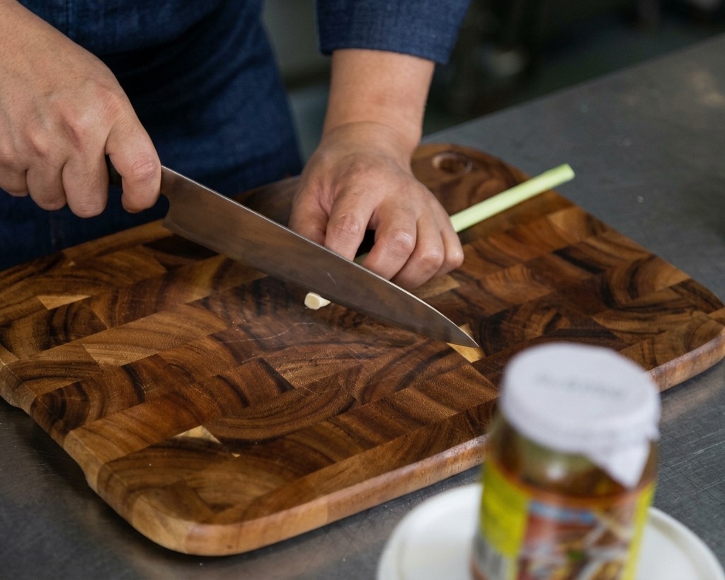 Manos usando un cuchillo afilado para picar finamente tallos de citronela fresca sobre una tabla de madera.
