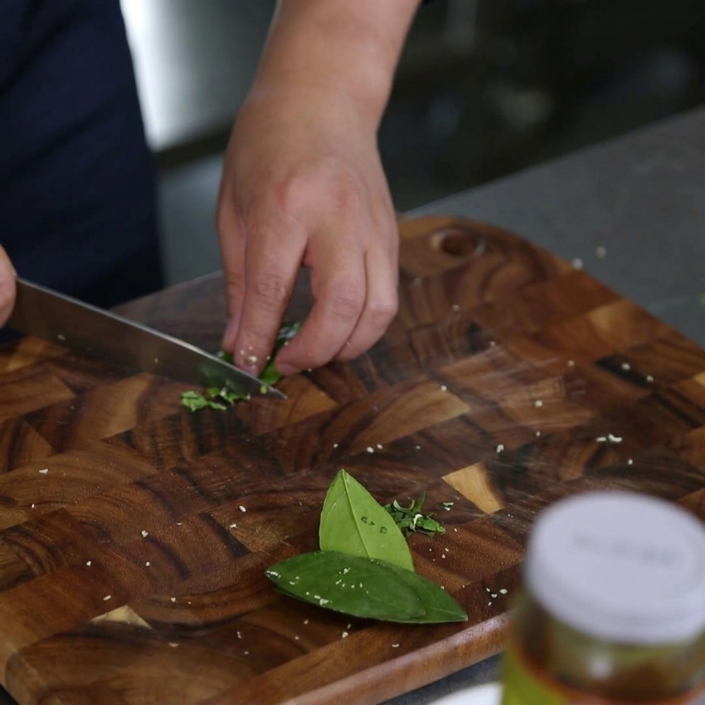 Primer plano de manos usando un cuchillo de chef para quitar el nervio central de hojas de lima kaffir en una tabla de cortar.