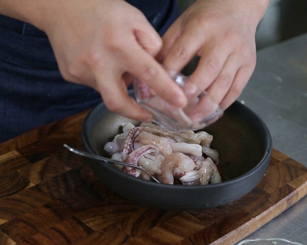 Manos vertiendo calamar crudo en rodajas y camarones en un bol de mezcla con salsa y otros mariscos.
