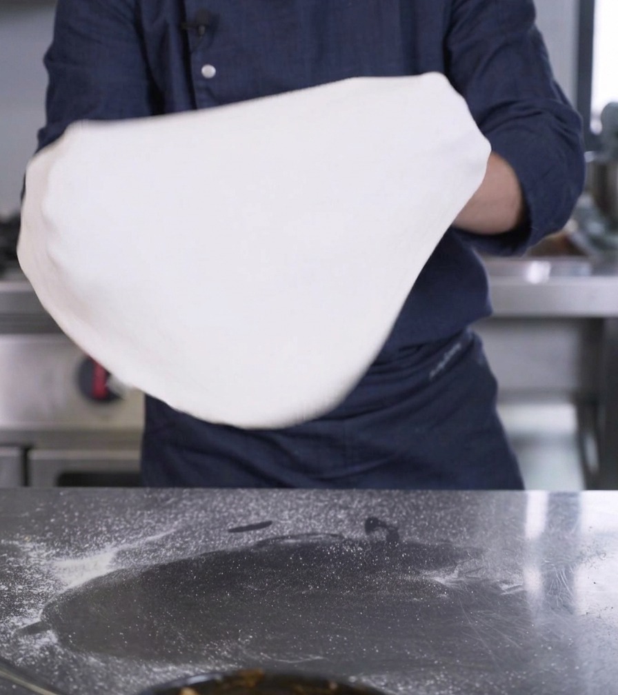 Manos de una persona sosteniendo y estirando un disco grande y fino de masa de pizza blanca.