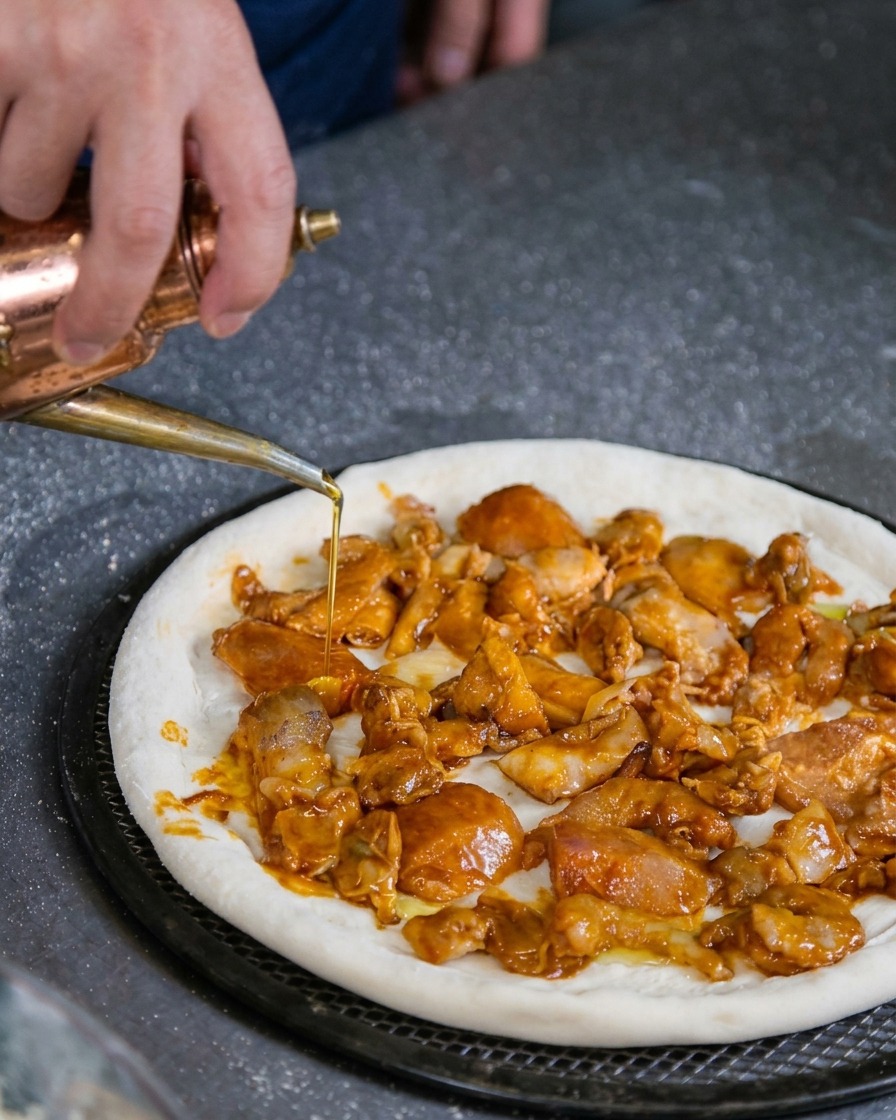Una mano usando un dispensador de aceite de cobre para rociar aceite sobre una base de pizza cruda cubierta de mariscos.