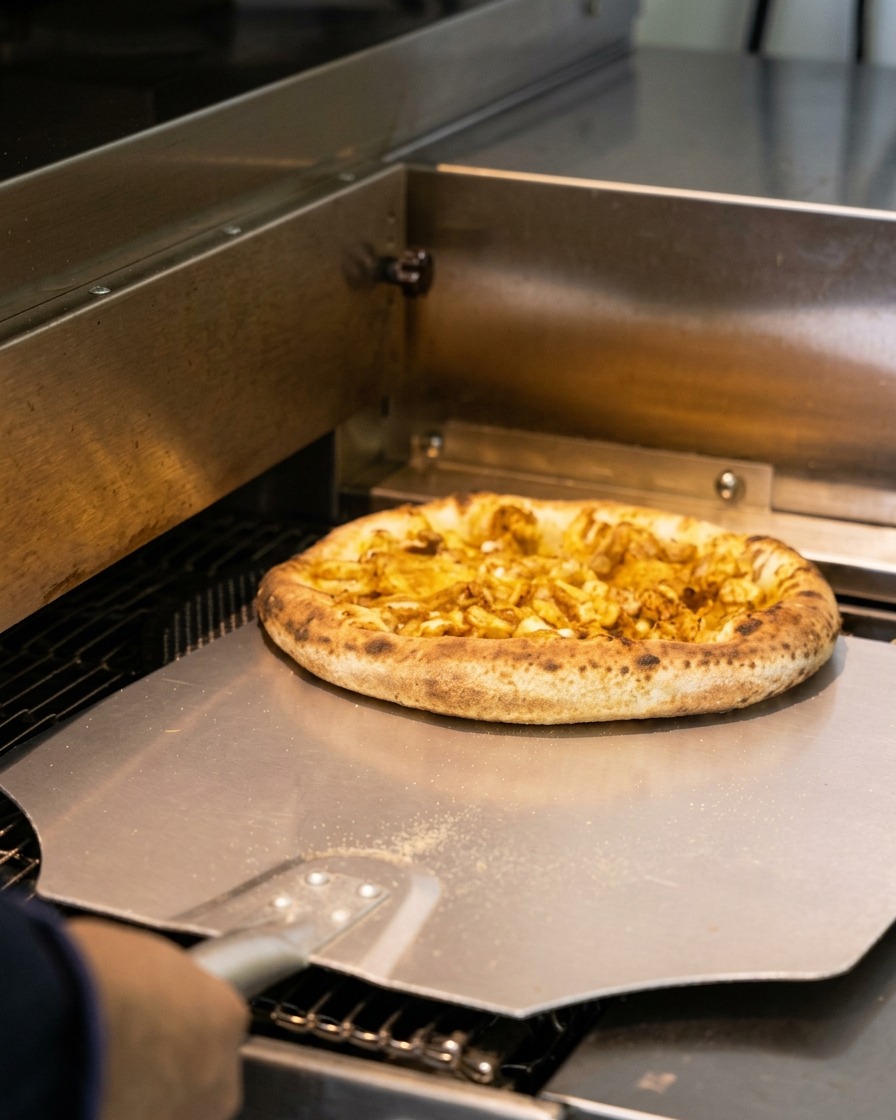 Una pizza de mariscos recién horneada con bordes tostados siendo retirada de un horno con una pala metálica.