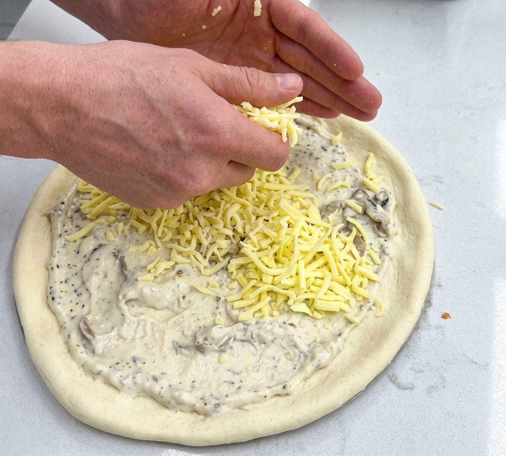 Dos manos esparciendo puñados de queso mozzarella rallado sobre una base de pizza cubierta de salsa blanca.