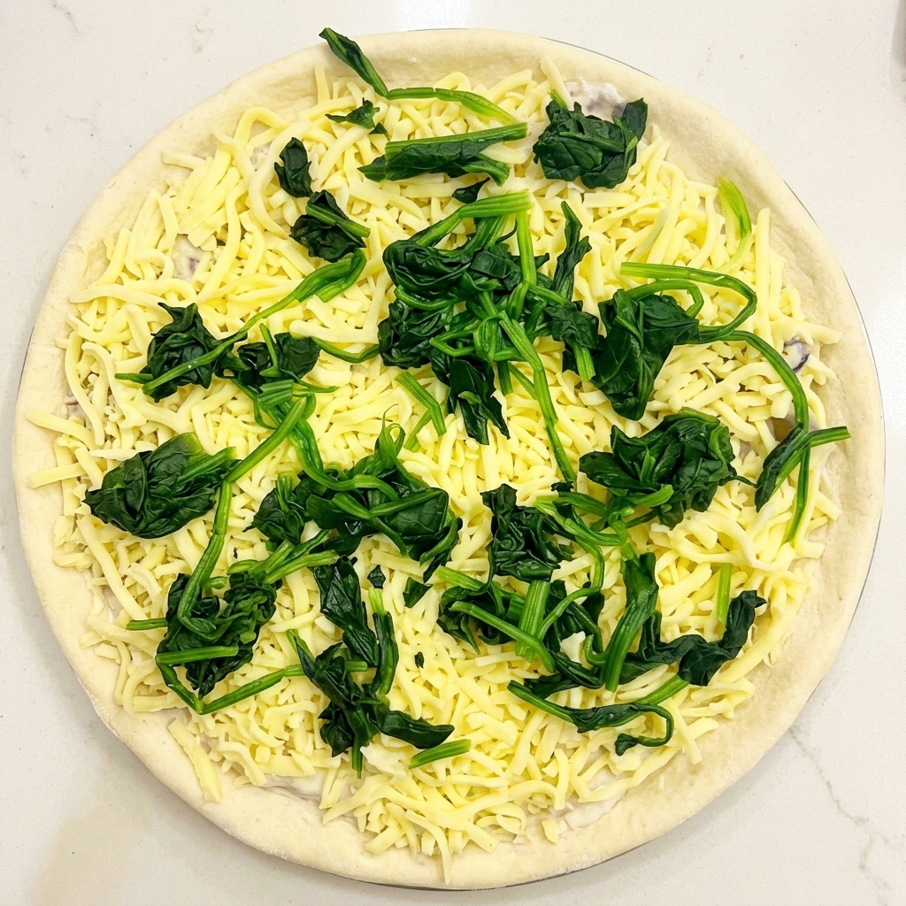 Vista superior de masa de pizza con salsa, mozzarella y porciones distribuidas de espinacas verdes escaldadas.