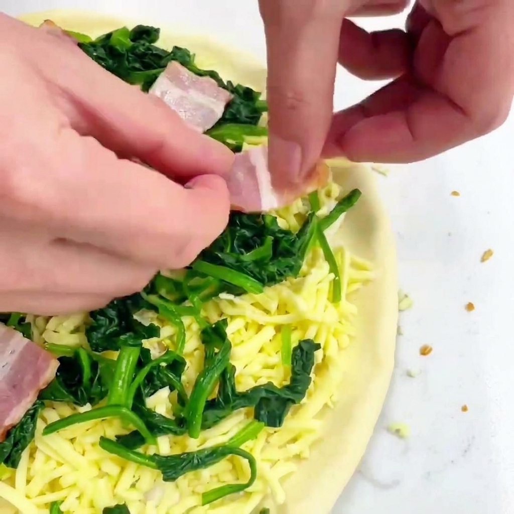 Manos colocando cuadrados de bacon crudo de color rosado sobre una pizza con queso y espinacas verdes.