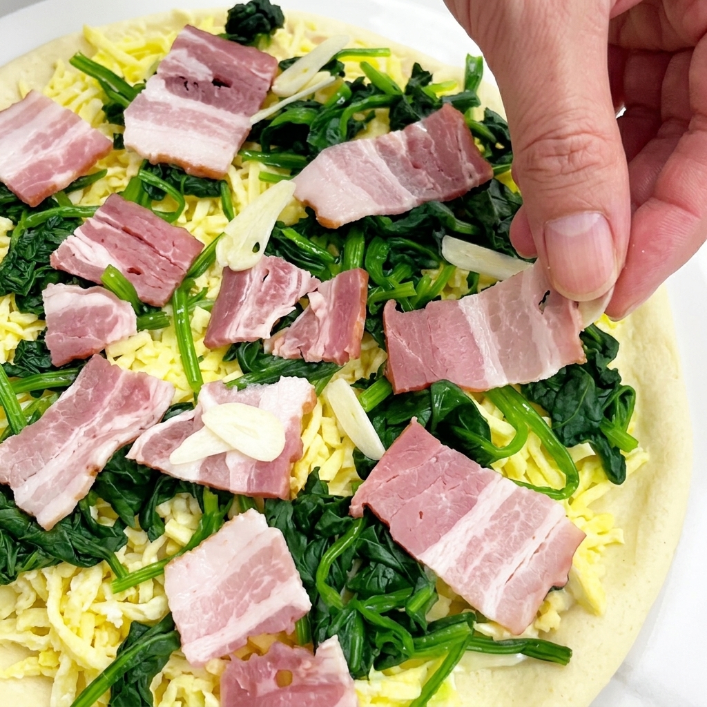 Mano dejando caer finas láminas de ajo crudo sobre una pizza cubierta con queso, espinacas y cuadrados de bacon.