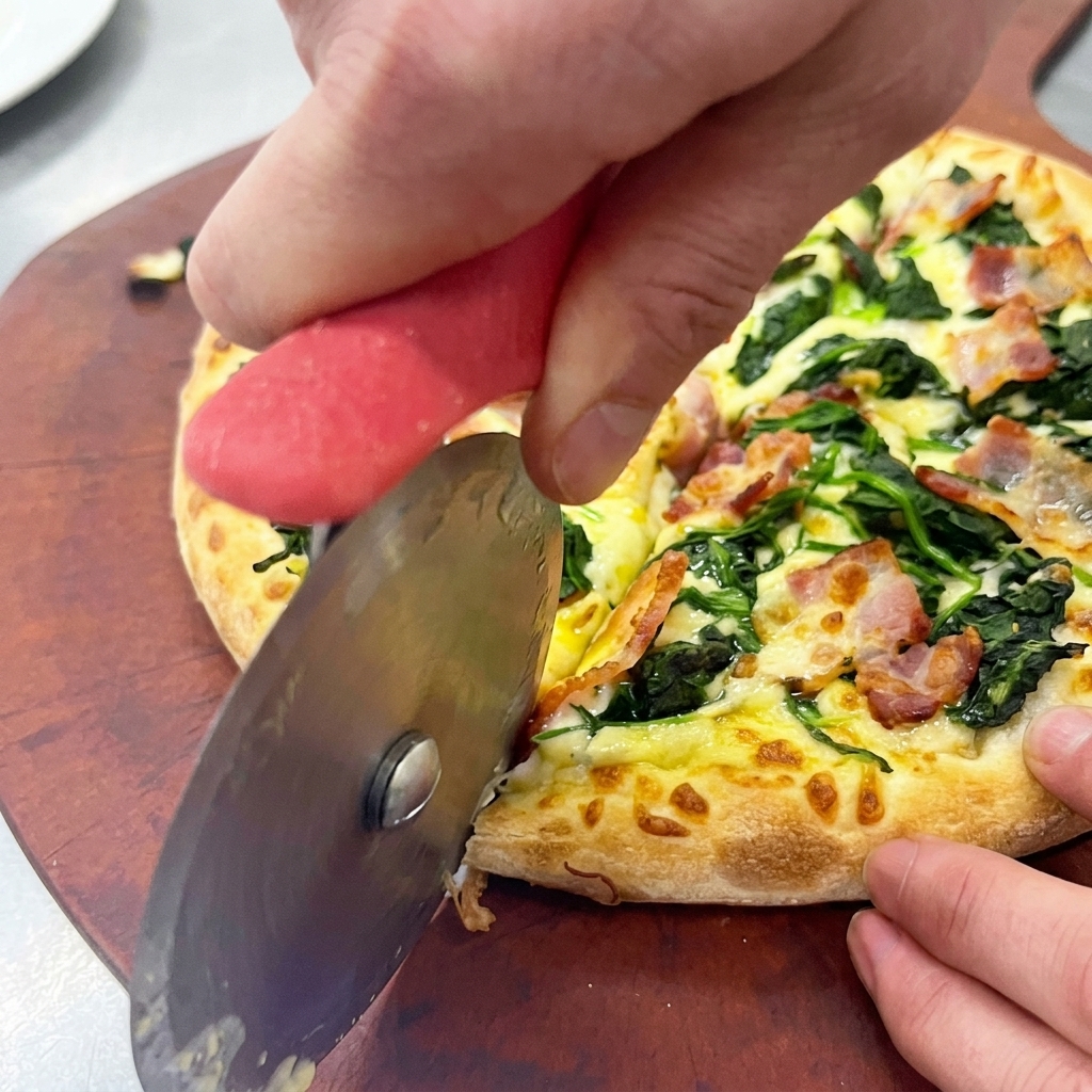 Manos utilizando un cortador de pizza circular para trocear una pizza de espinacas y bacon sobre una tabla de madera.