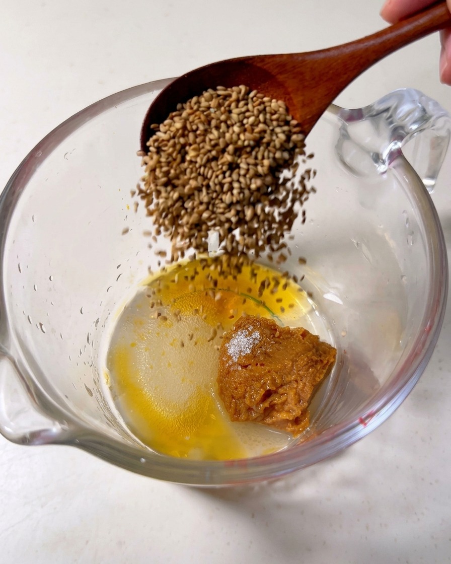 Una cuchara de madera vertiendo semillas de sésamo tostadas en una jarra con pasta de miso, azúcar y aceite de sésamo.