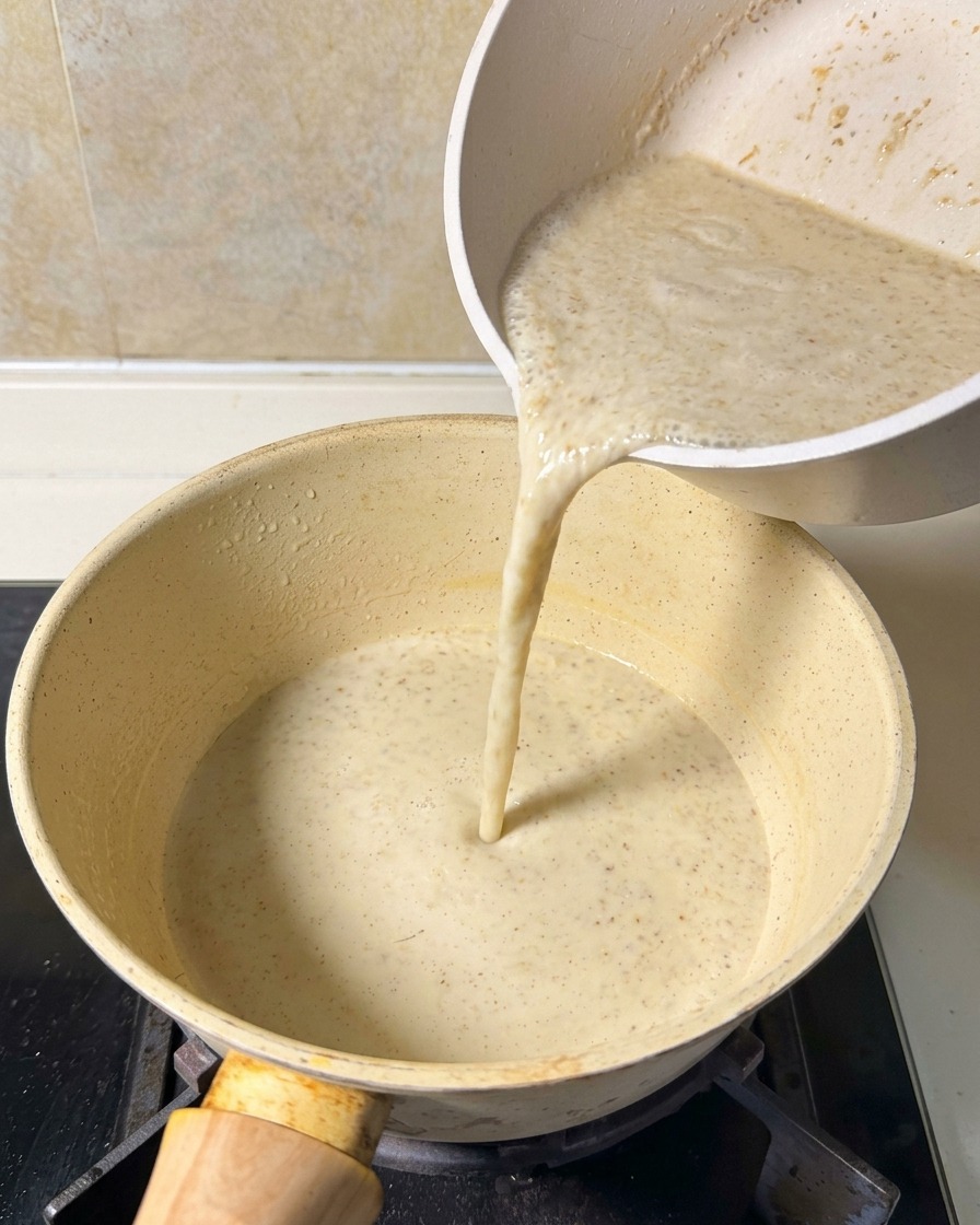 Una mezcla de sopa beige y suave vertiéndose desde una jarra blanca a una olla pequeña sobre el fuego.
