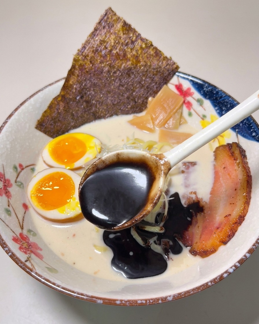 Una cuchara vertiendo aceite de ajo negro espeso y oscuro sobre un cuenco de ramen terminado.