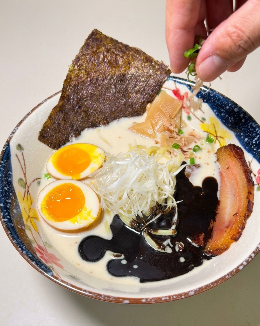 Una mano espolvoreando cebolleta picada sobre un cuenco de ramen con aceite de ajo negro, chashu y huevo meloso.