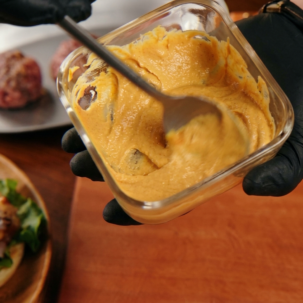 Una mano mezclando una salsa de hamburguesa de color naranja cremoso con una cuchara en un recipiente de cristal.