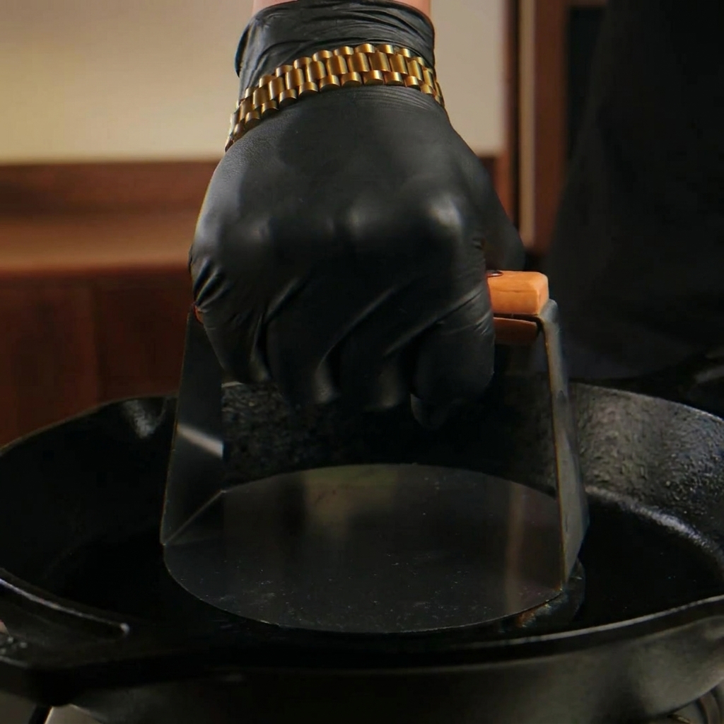 Una mano con guante negro usa una prensa metálica pesada para aplastar una bola de carne cruda sobre una sartén de hierro humeante.