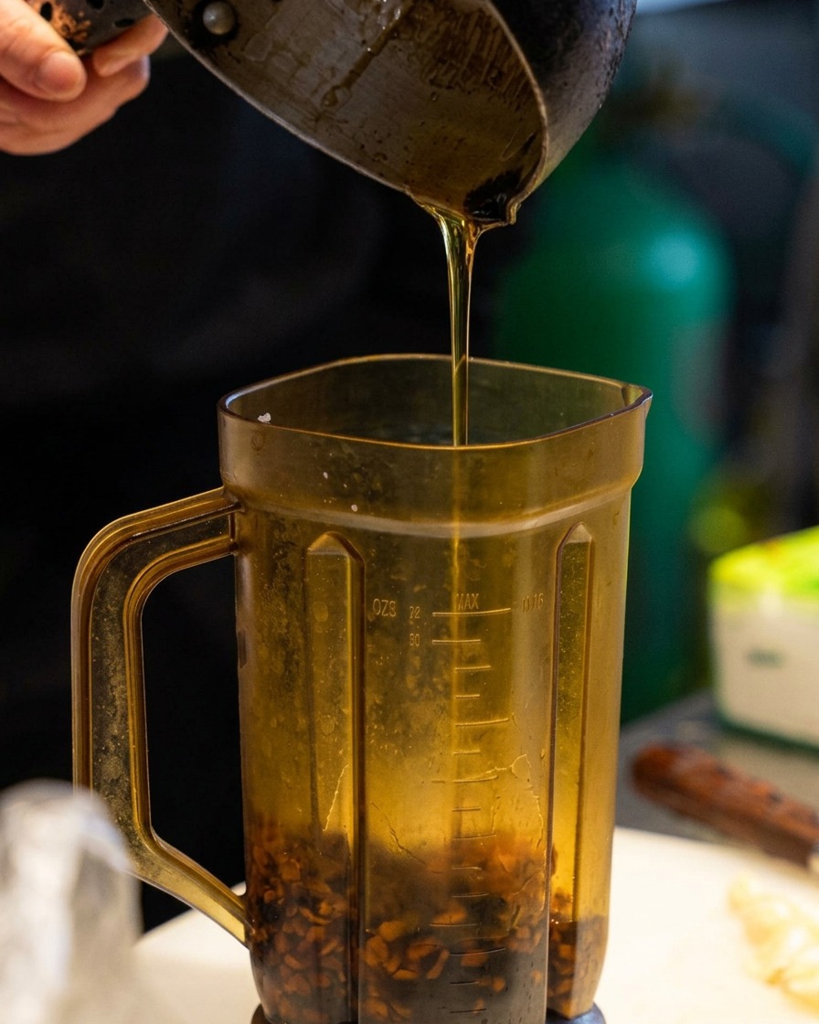 Vertiendo aceite de sésamo dorado desde una pequeña jarra metálica al vaso de la licuadora que contiene trozos de ajo frito oscuro.