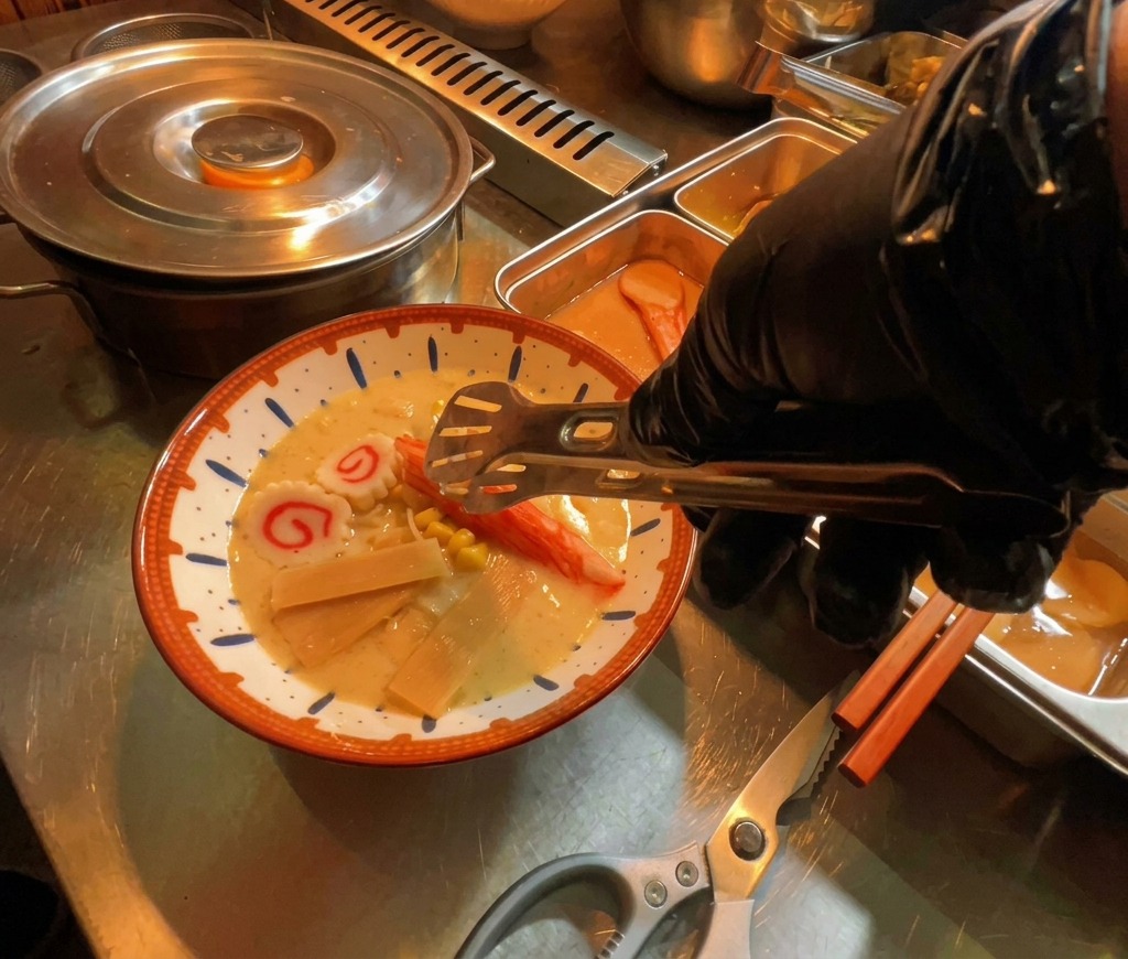 Una mano con guante colocando rodajas de narutomaki en un humeante bol de ramen.