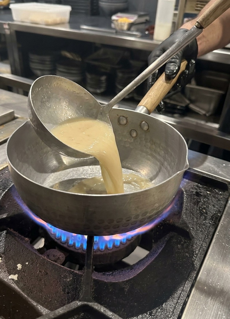 Un cucharón de metal vertiendo caldo de cerdo claro en una olla sobre una estufa de gas.