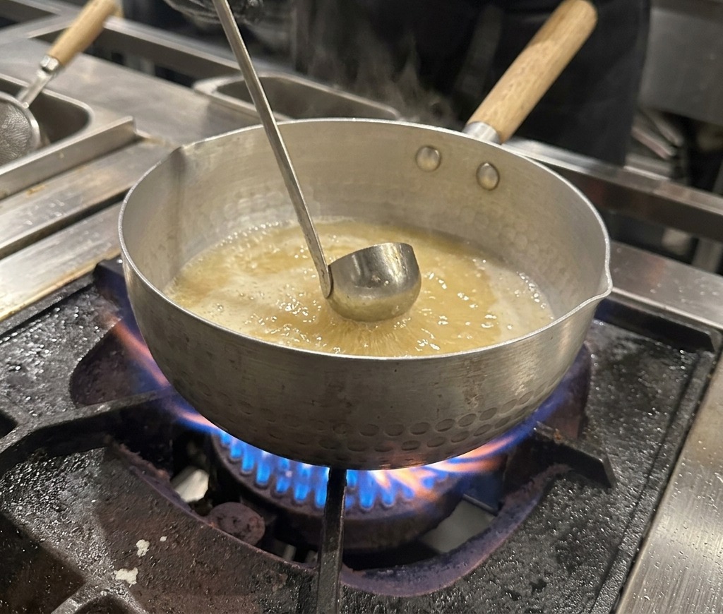 Una mano con guante sosteniendo un bol de metal y añadiendo salsa de soja oscura a una olla de caldo hirviendo.