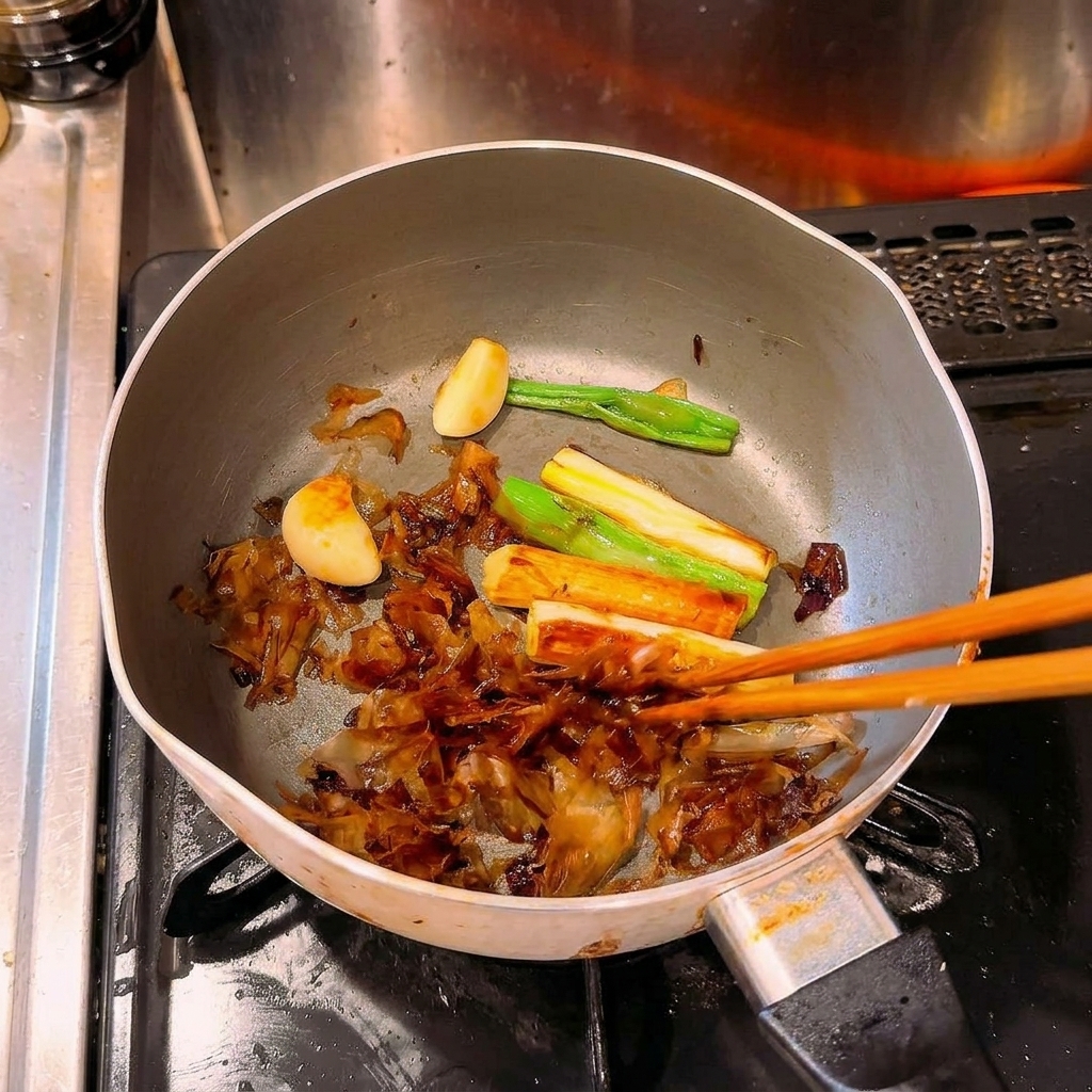 Palillos removiendo cebolletas, ajos y copos de bonito muy caramelizados y oscuros en una olla.