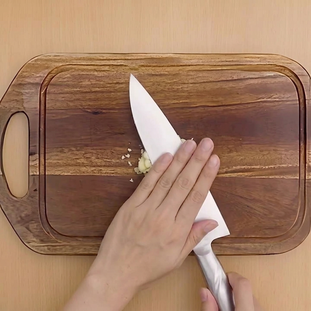 Una mano presionando el lateral de un cuchillo de chef para aplastar un diente de ajo sobre una tabla de cortar.