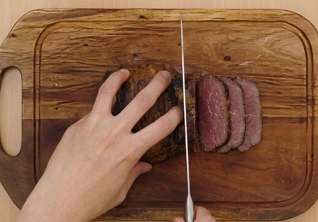 Manos sujetando el rollo de cerdo mientras un cuchillo afilado corta rodajas finas sobre una tabla de madera.
