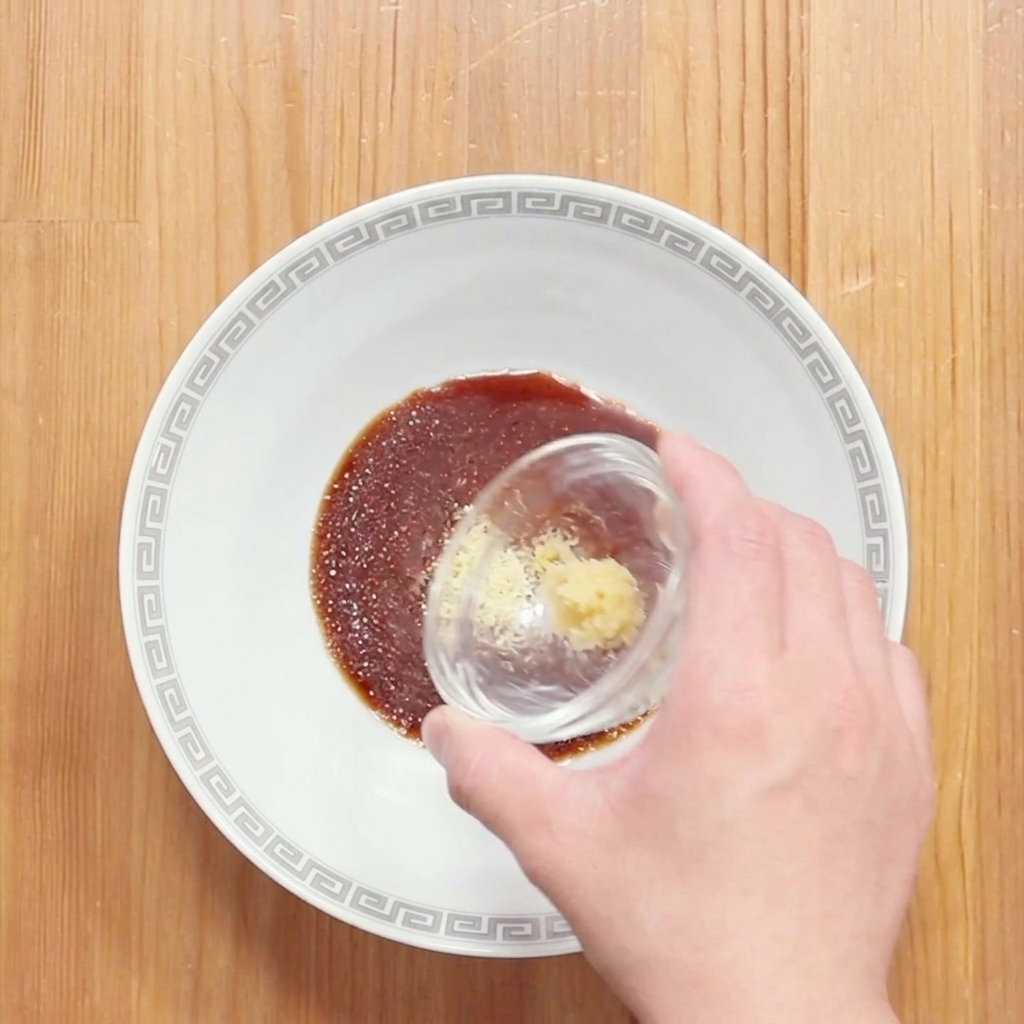 Una mano añadiendo jengibre rallado a un tazón con la mezcla oscura de salsa de soja para el ramen.