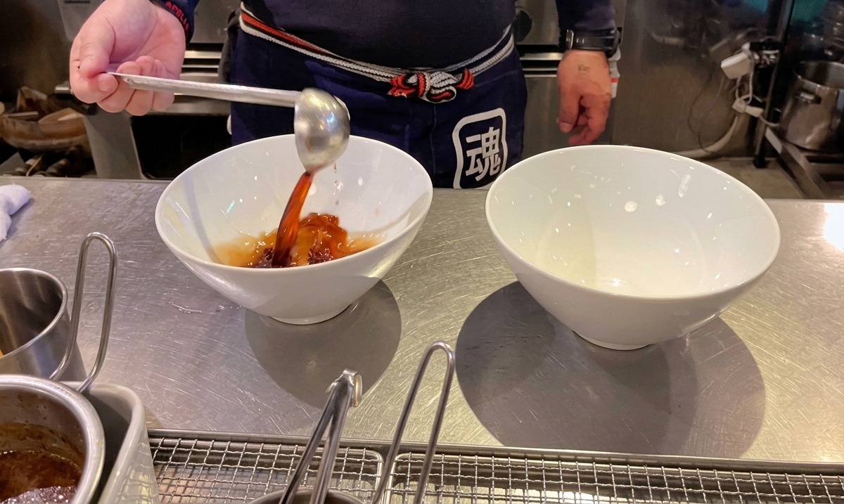 Cocinero vertiendo tare de salsa de soja oscura en cuencos de ramen blancos.