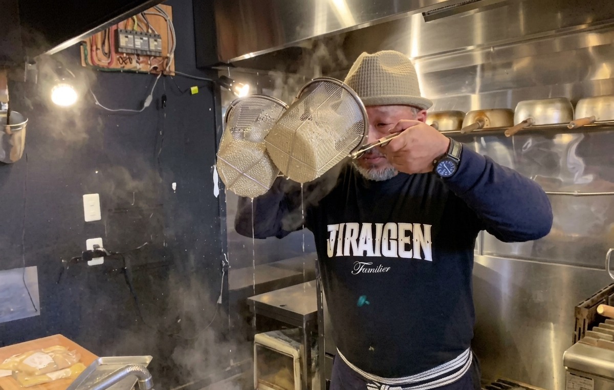 Cocinero agitando enérgicamente los coladores de fideos para eliminar el agua del ramen cocido.