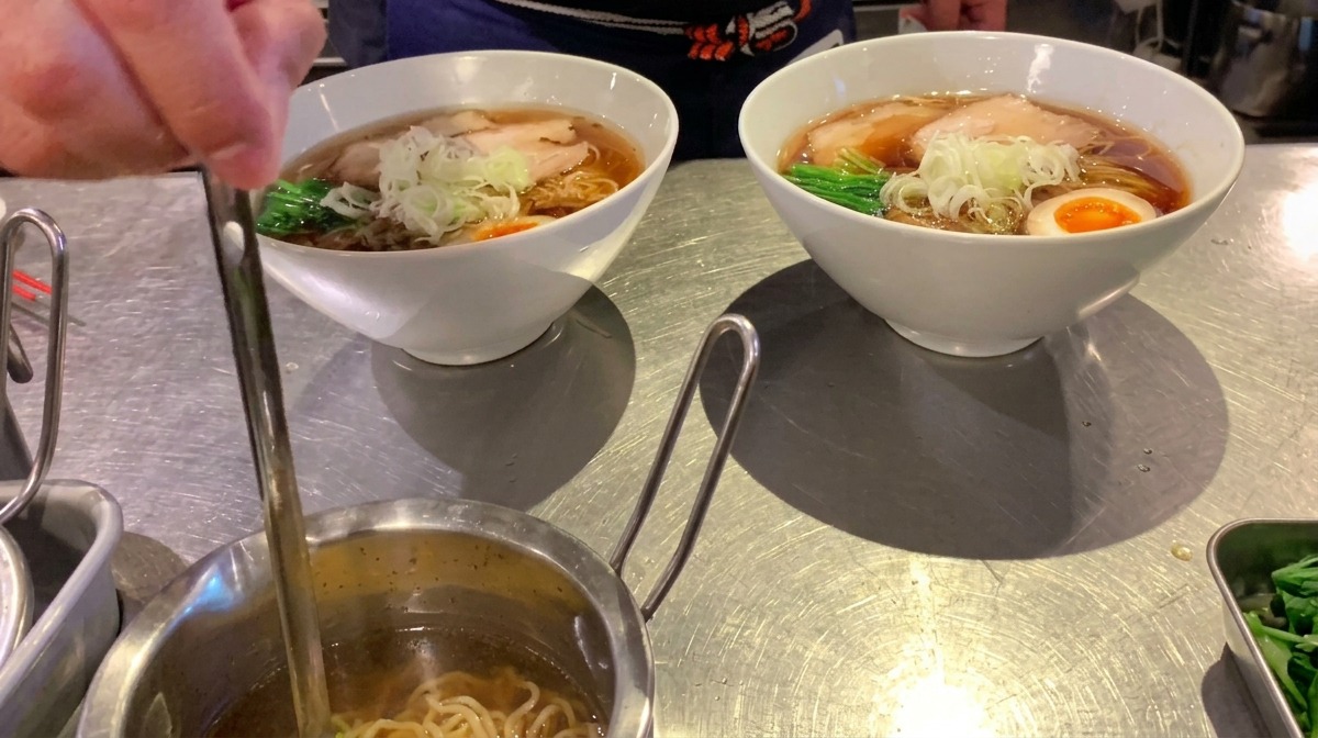 Un pequeño cazo rociando aceite aromático transparente sobre un cuenco de ramen terminado.