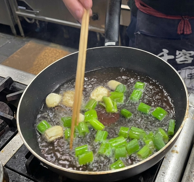 Friendo trozos grandes de cebolleta, dientes de ajo y rodajas de jengibre en la grasa de cerdo en una sartén oscura.