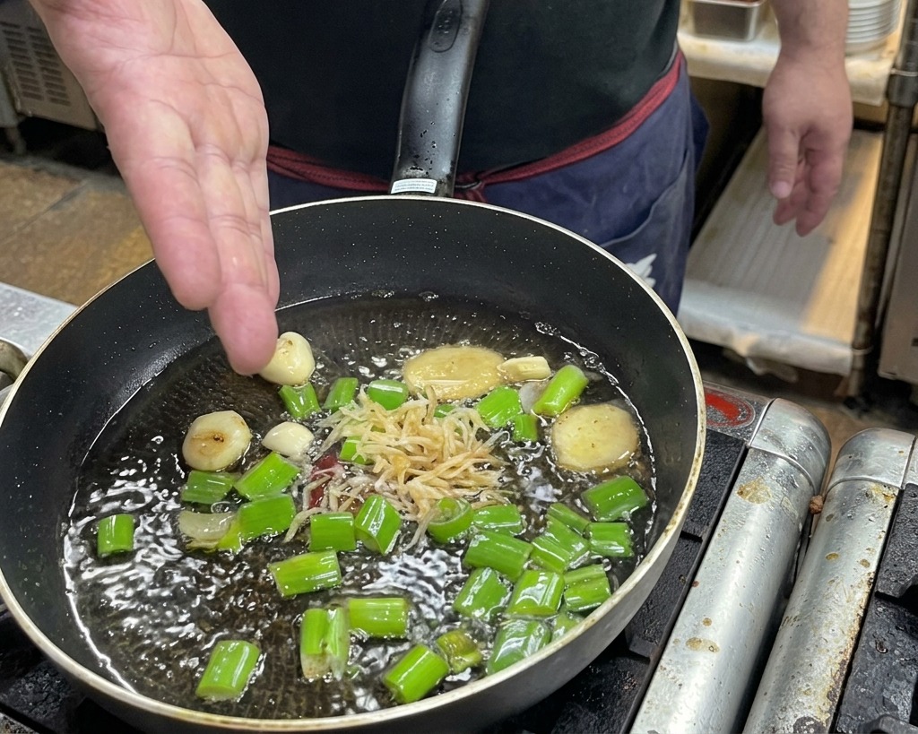 Una mano echando camarones secos a una sartén con aceite caliente, jengibre, ajo y cebolleta.