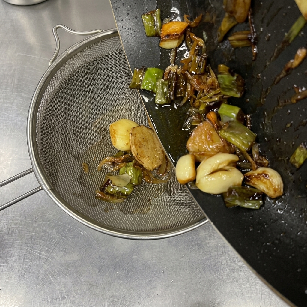 Vertiendo aceite caliente y aromáticos dorados como ajo y cebolleta desde una sartén a un colador fino.