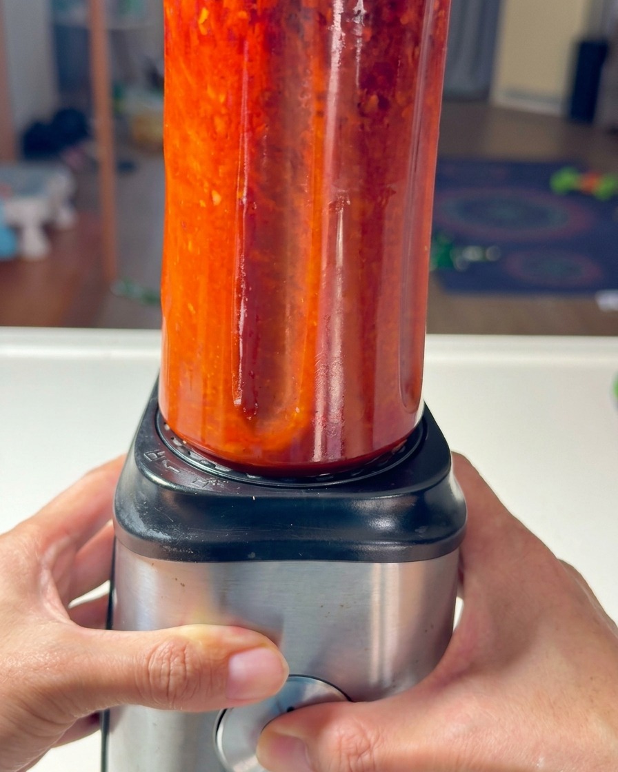 Manos asegurando un vaso de batidora lleno de pasta de chile roja en una base metálica.