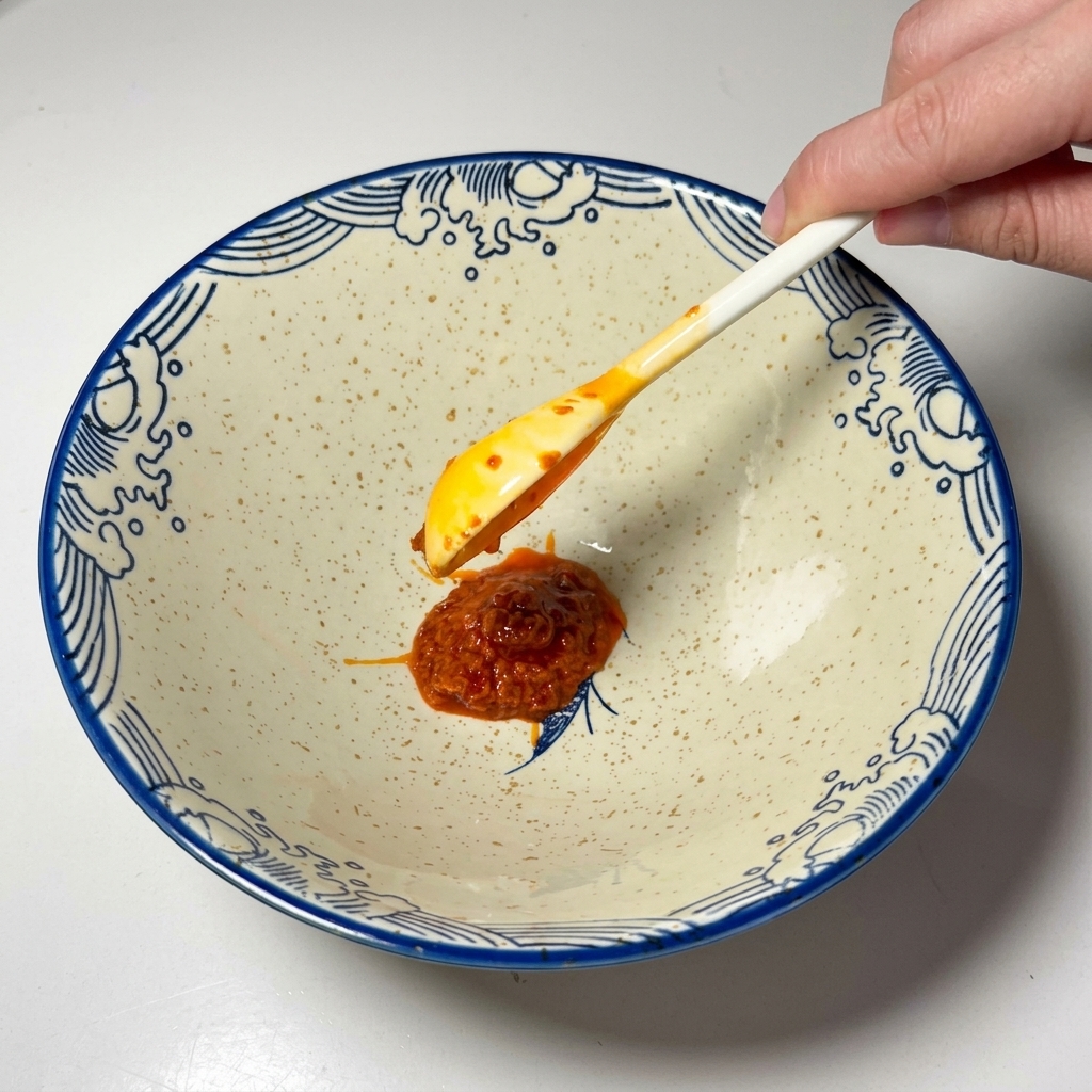 Una cucharada de pasta de chile roja colocándose en el fondo de un cuenco cerámico.