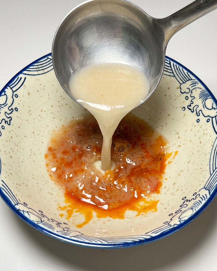 Caldo Tonkotsu blanco y cremoso vertiéndose desde un cazo sobre la pasta roja.