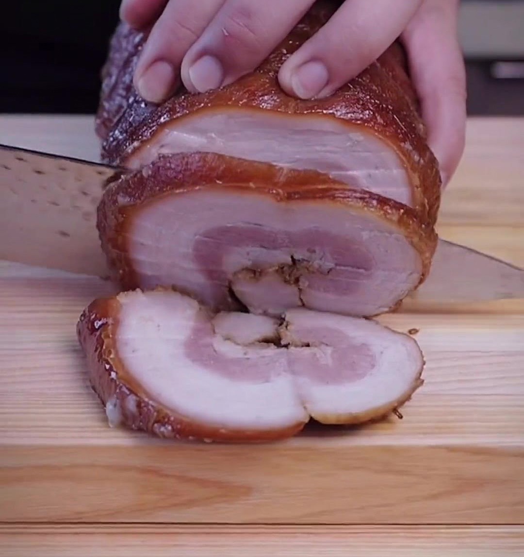 Manos con un cuchillo afilado cortando rodajas gruesas de un rollo de chashu frío sobre una tabla de madera.