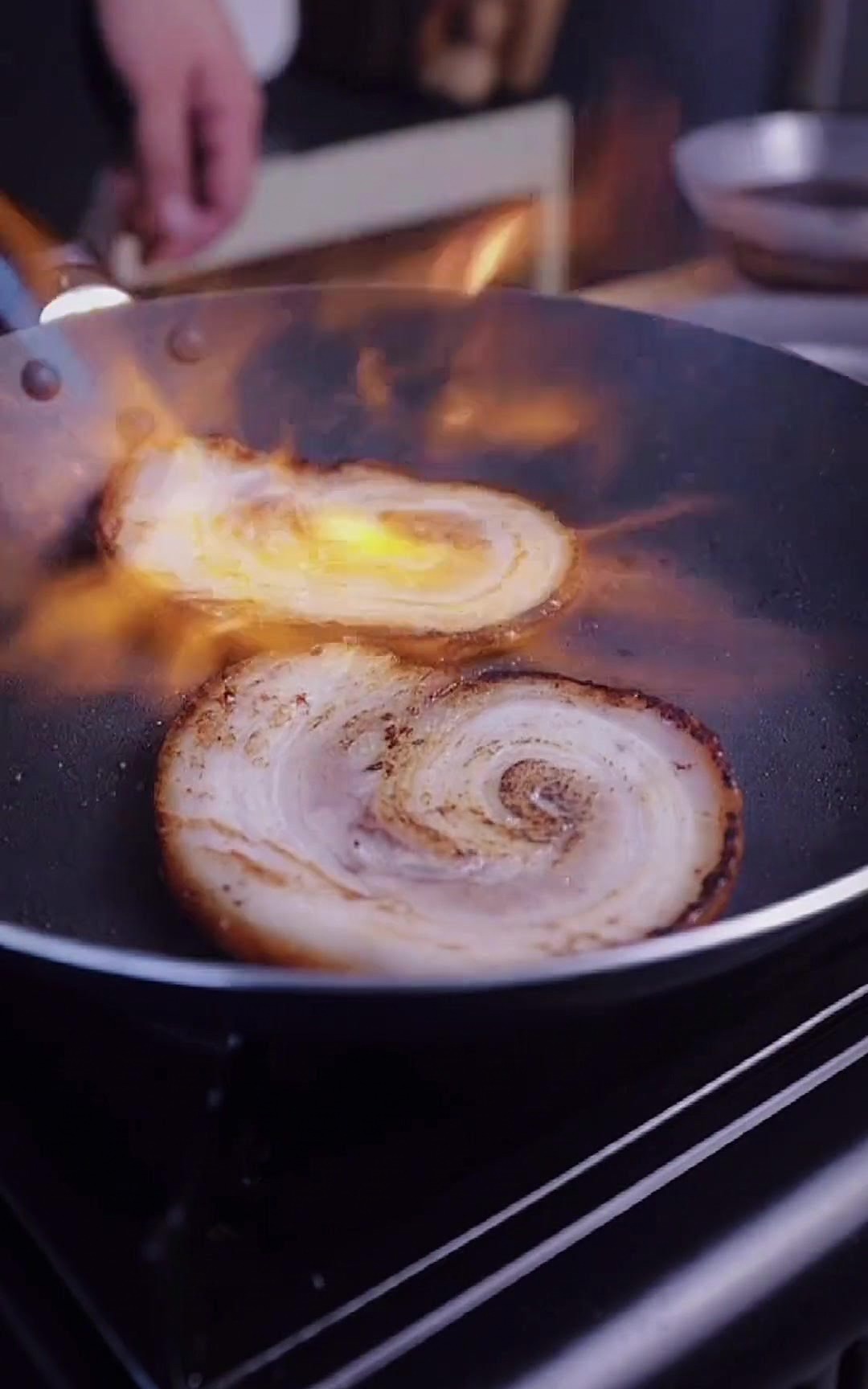 Dos rodajas de chashu en una sartén recibiendo la llama naranja de un soplete de cocina.