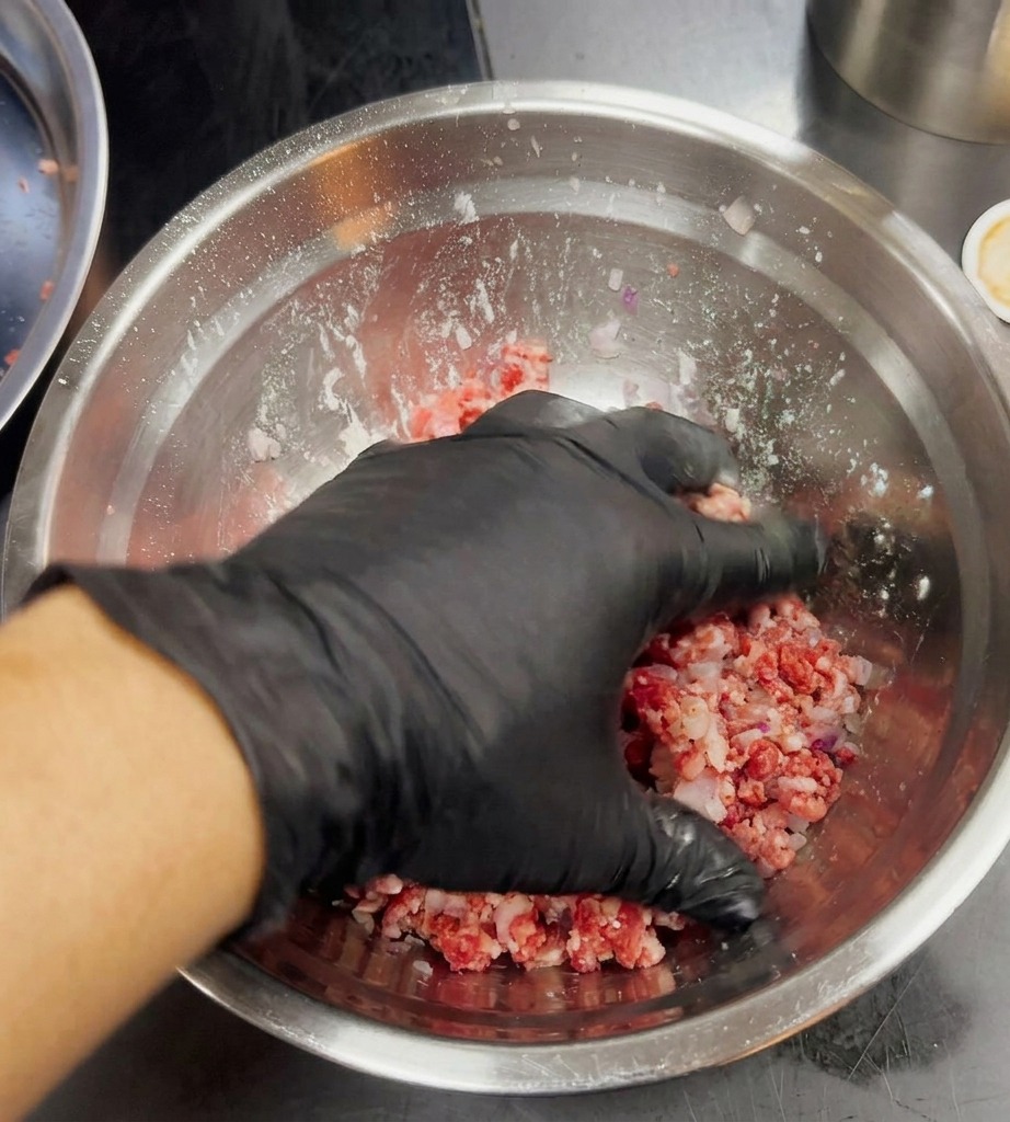Una mano con guante negro mezclando ligeramente la carne picada y la cebolla morada en un bol plateado.
