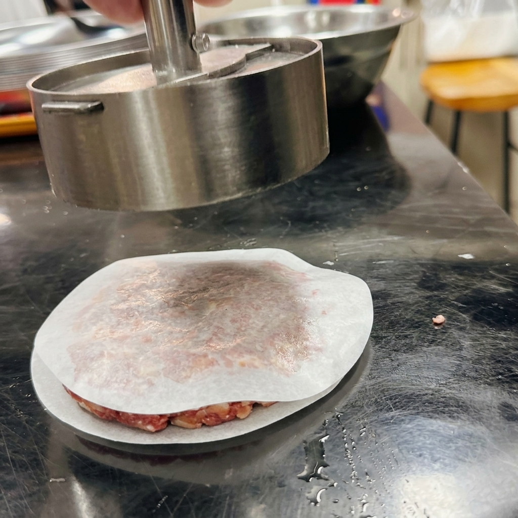 Una prensa de hamburguesas metálica cilíndrica dando forma a la carne sobre un trozo de papel de horno.