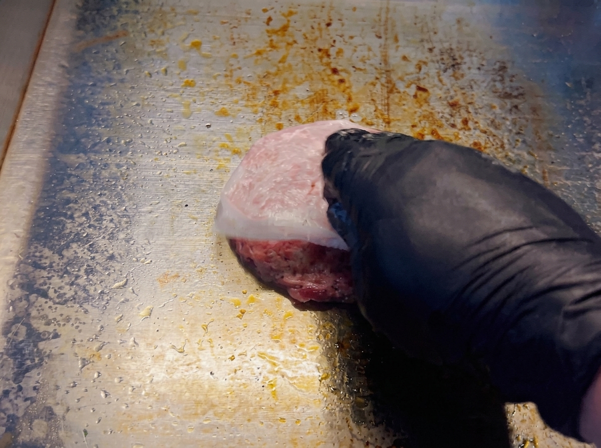 Una mano con guante retirando el papel de horno de una hamburguesa de ternera cruda sobre una plancha caliente.