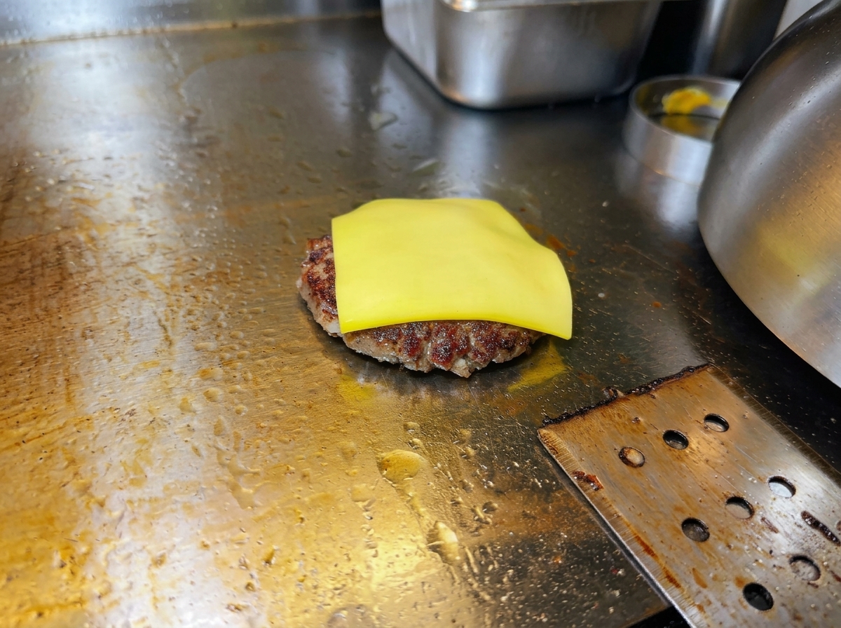 Una loncha de queso amarillo empezando a fundirse sobre una hamburguesa sellada en la plancha.