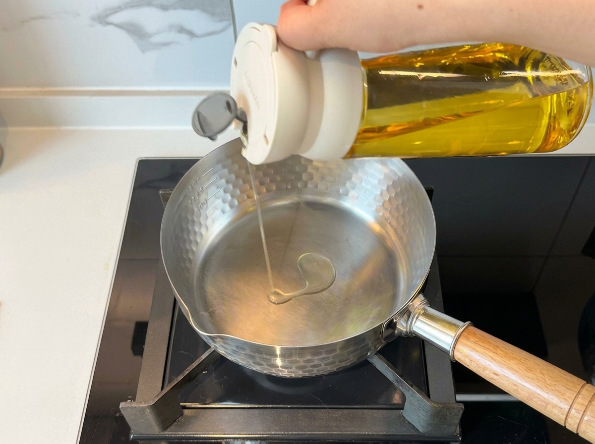 Una mano vertiendo aceite de cocina desde una botella en una olla de acero inoxidable limpia sobre el fuego.
