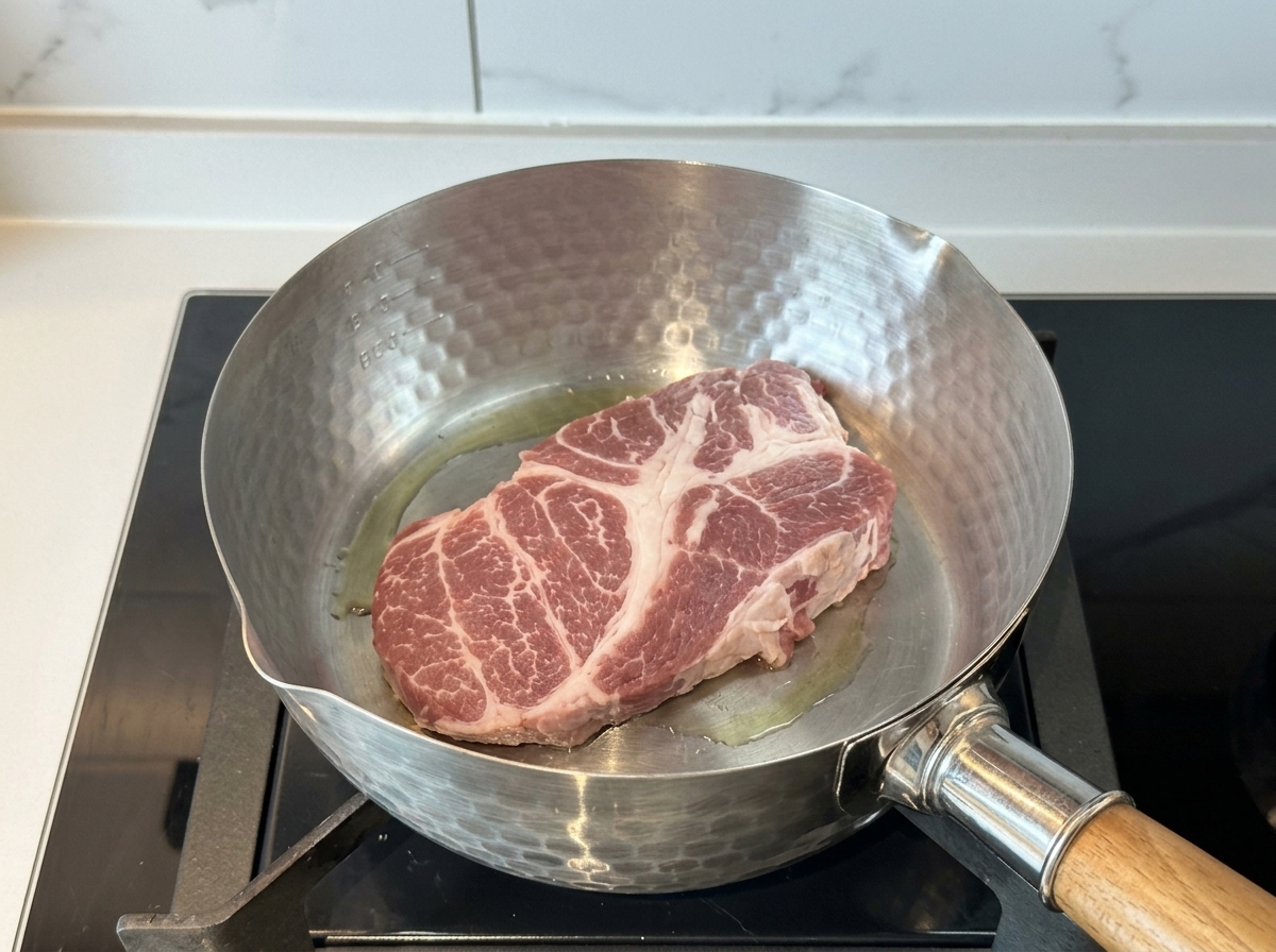 Un trozo grueso de aguja de cerdo cruda sellándose en una olla de acero inoxidable caliente con un poco de aceite.