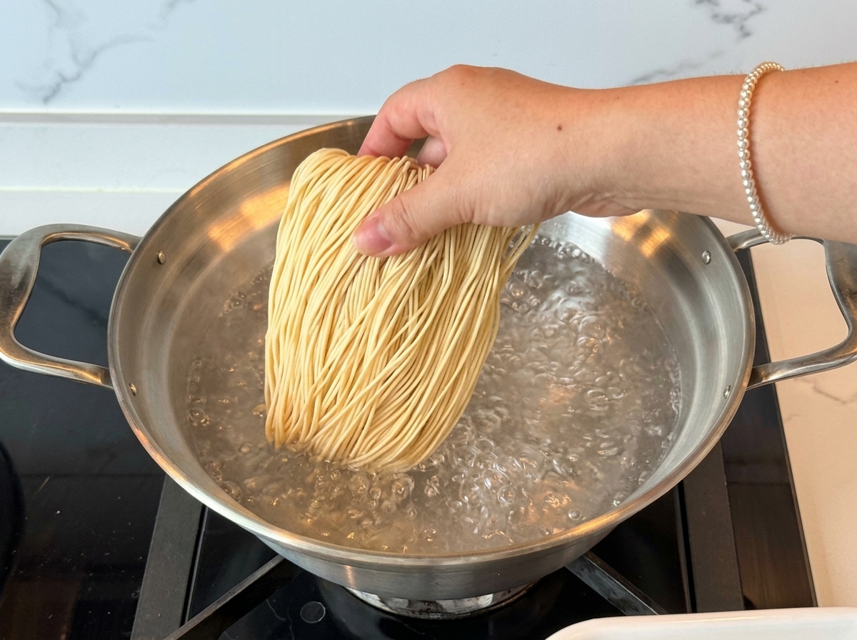 Una mano añadiendo un nido de fideos ramen secos en una olla con agua hirviendo con fuerza.