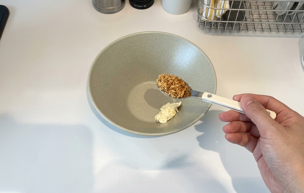 Una mano con una cuchara de ajo frito crujiente sobre un bol cerámico que contiene una porción de mayonesa.