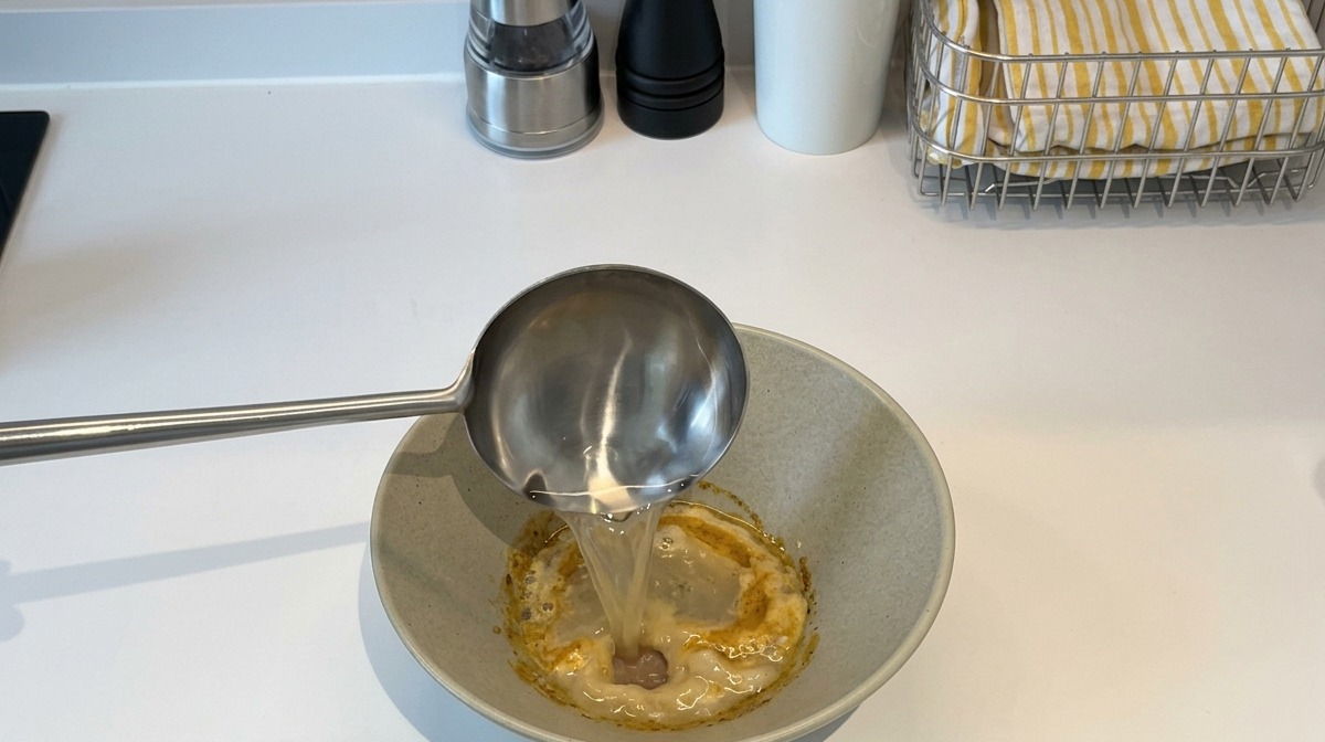 Un cazo de acero inoxidable vertiendo agua caliente en un bol que contiene la pasta espesa de condimentos.