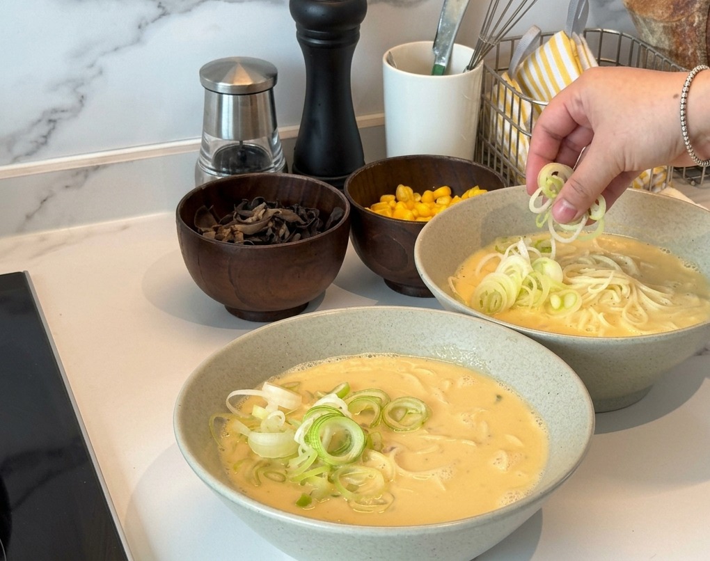 Una mano espolvoreando cebolleta verde finamente picada sobre dos boles de ramen cremoso.