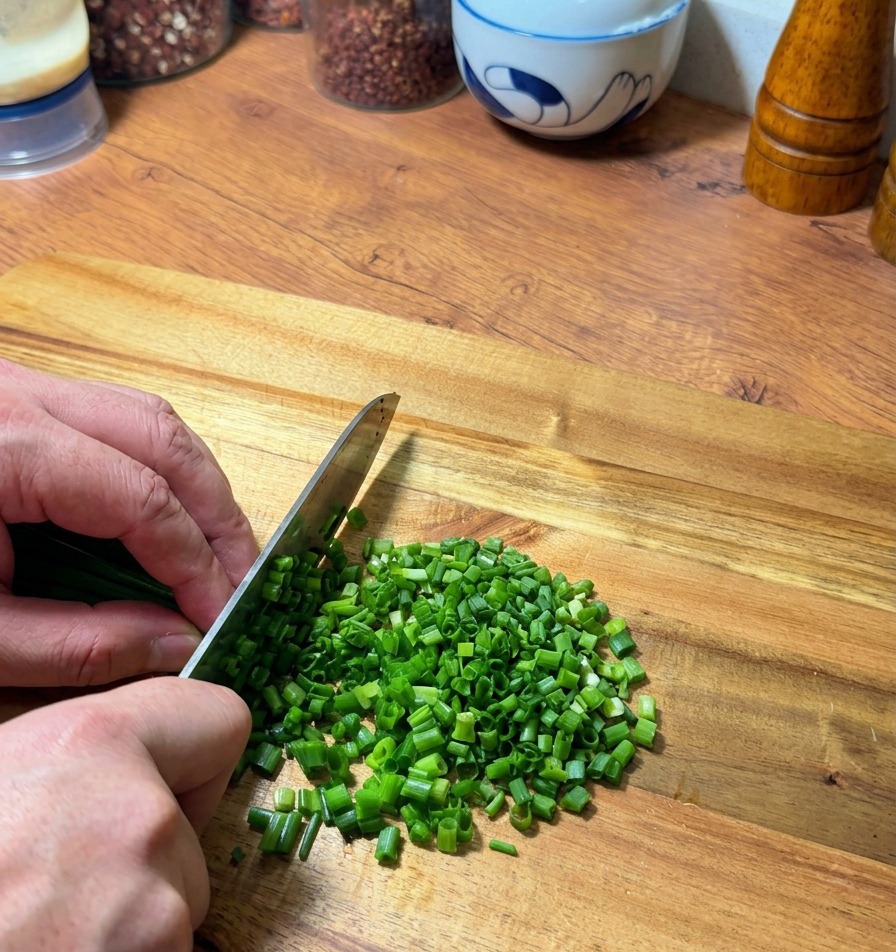 Manos picando cuidadosamente cebolletas verdes frescas sobre una tabla de cortar de madera con un cuchillo.