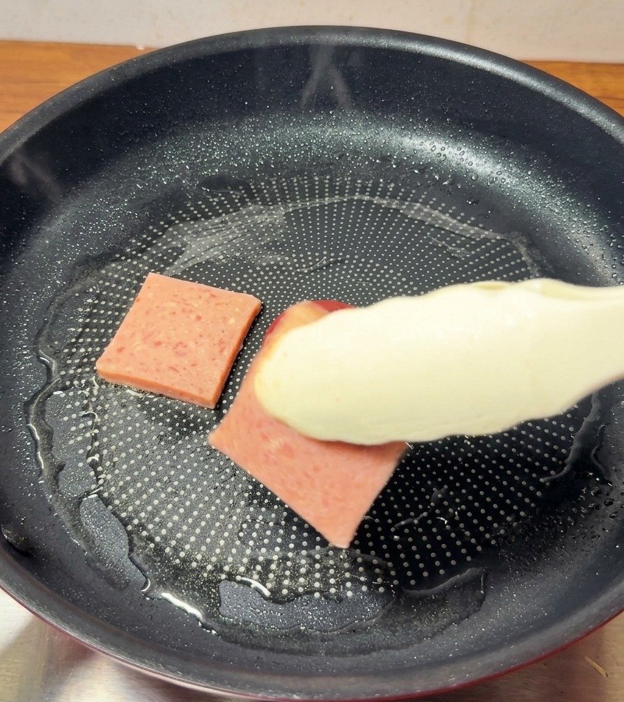 Lonchas de carne de almuerzo dorándose en una sartén antiadherente, con unas pinzas dando la vuelta a una pieza para mostrar la costra dorada.
