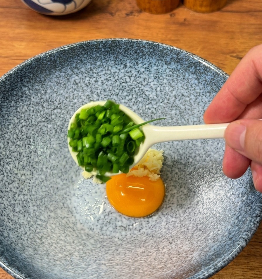 Una cucharada de cebolletas picadas recién cortadas añadiéndose a un bol de servicio que contiene ajo picado y una yema de huevo cruda.