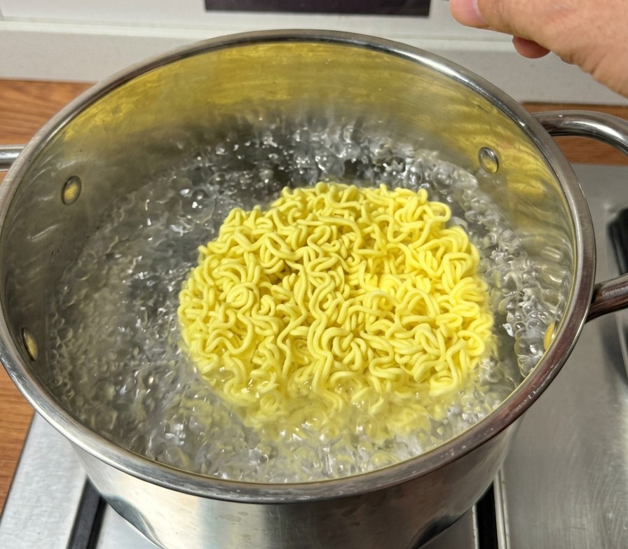 Un bloque de fideos ramen instantáneos amarillos y secos cayendo en una olla de agua hirviendo.